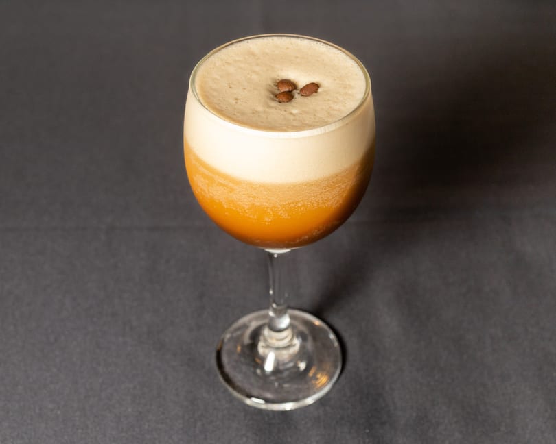 Espresso martini in a stemmed glass, topped with foam and three coffee beans on a dark surface.