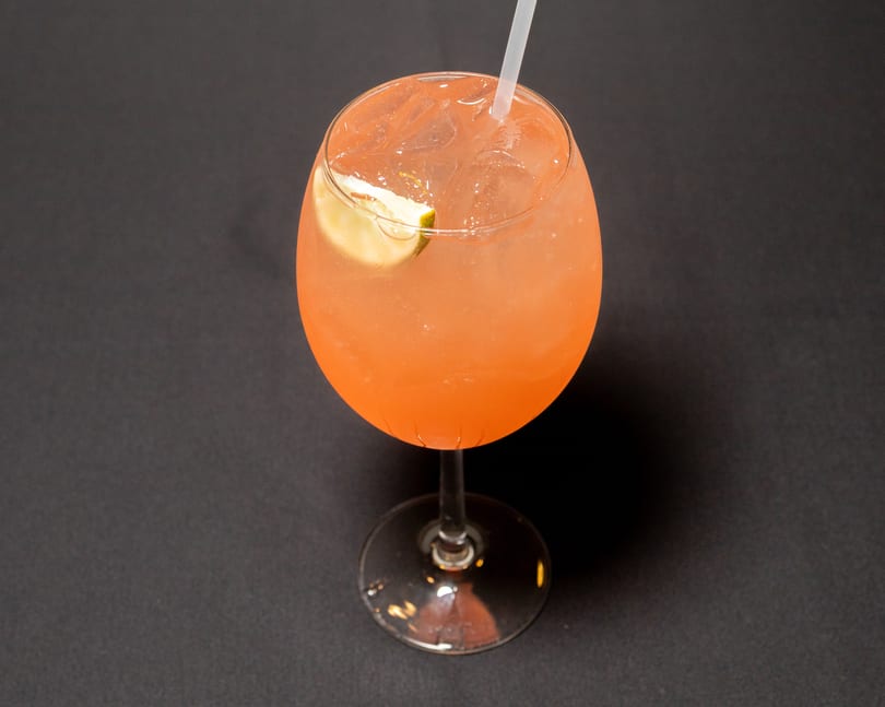 A vibrant orange cocktail with ice and a lime wedge, served in a stemmed glass on a dark surface.