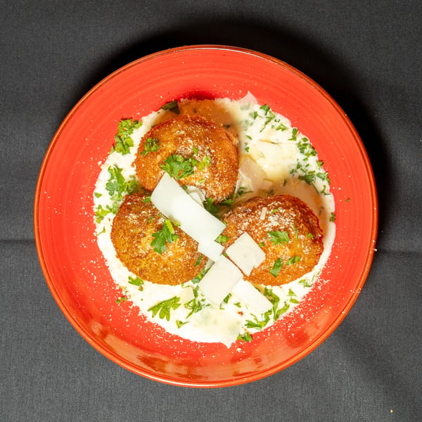 Three golden-brown arancini balls in a creamy sauce, garnished with parsley and parmesan shavings on a red plate.