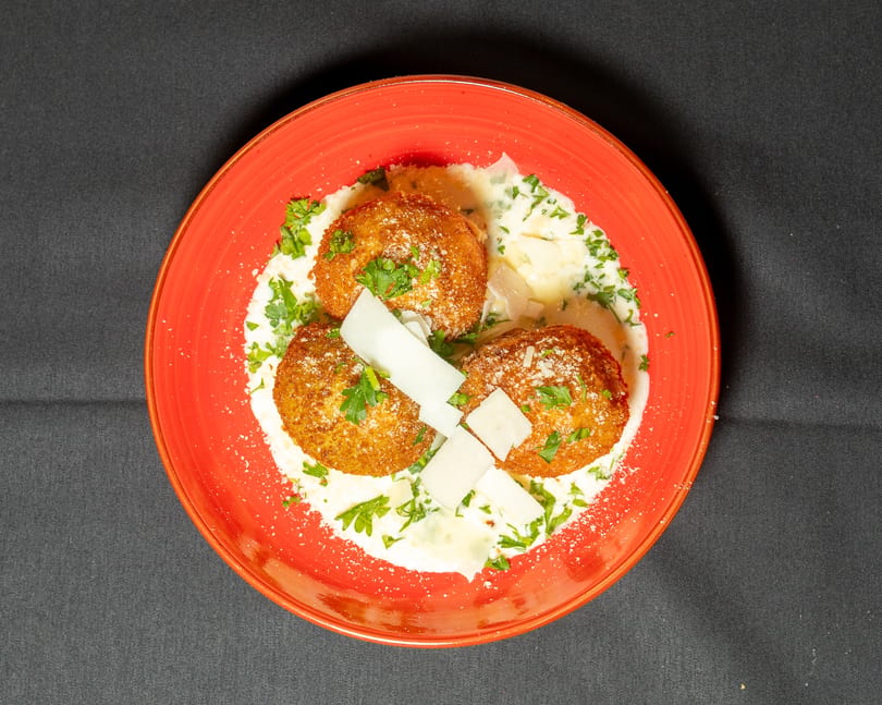 Three golden-brown arancini balls in a creamy sauce, garnished with parsley and parmesan shavings on a red plate.