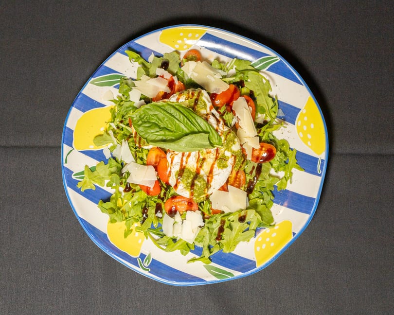Burrata salad with arugula, cherry tomatoes, pesto, balsamic glaze, and parmesan on a striped plate.