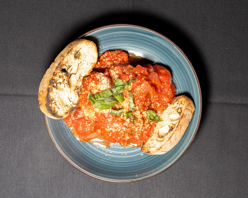 Meatballs in red sauce with parmesan cheese, basil, and toasted bread on a blue plate.